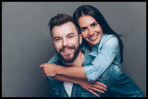 Handsome young man piggybacking beautiful woman and smiling while standing against grey background
